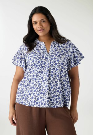 Donna che indossa una camicetta bianca con stampa floreale blu e maniche svolazzanti, abbinata a pantaloni marroni larghi, in piedi davanti a uno sfondo semplice.