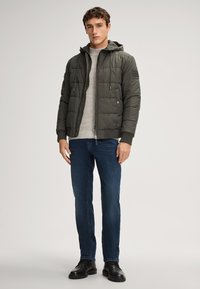 Giacca imbottita verde oliva con cappuccio, zip frontale e polsini a coste, abbinata a un maglione grigio chiaro e jeans scuri, completata da stivaletti neri.