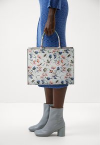Torebka tote w kwiatowy wzór z jasnoniebieskim tłem, o wyraźnej formie, z podwójnymi uchwytami u góry i szarymi detalami. Nosi ją osoba w teksturowanej niebieskiej sukience i szarych butach.