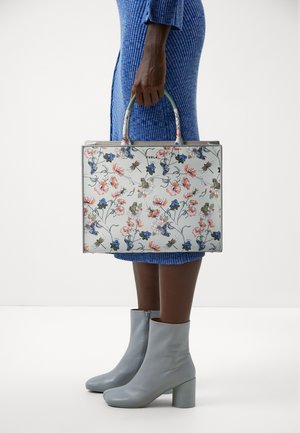 Bloemmotief schoudertas met een lichtblauwe achtergrond, gestructureerde vorm, dubbele handvatten bovenaan en grijze details. Gedragen met een textuur blauw jurk en grijze laarzen.