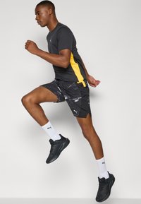 Schwarzers T-Shirt mit kurzen Ärmeln und gelbem Seitenstreifen; gemusterte schwarze Shorts; weiße Socken; schwarze Laufschuhe. Laufpose vor neutralem Hintergrund.