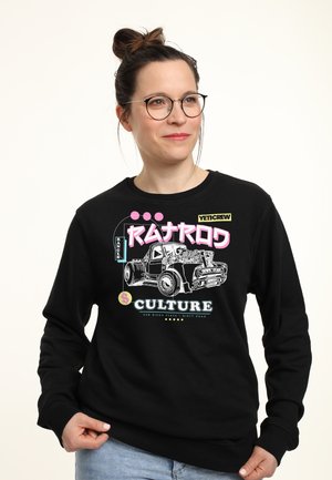 Kobieta w okularach, ubrana w czarną bluzę z nadrukiem przedstawiającym zabytkową ciężarówkę i kolorowy tekst "Ratrod Culture".