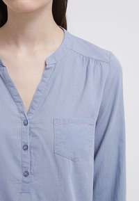 Chemise bleu clair à boutons avec col en V, une poche poitrine unique et de fines rayures verticales. Tissu doux avec une finition légèrement texturée.