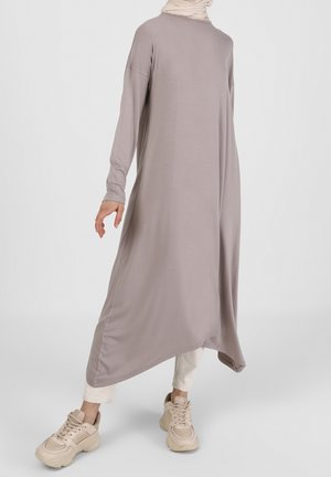 Femme portant une robe taupe ample à manches longues avec un ourlet asymétrique, un pantalon blanc, des baskets beiges et un foulard beige clair sur fond clair.