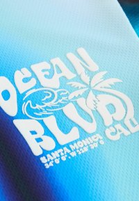 Tekstilpinta, jossa on vaaleansinisestä tummansiniseksi siirtyvä sävy. Siinä on valkoista kohotettua tekstiä "OCEAN BLVD" sekä palmupuugrafiikka.