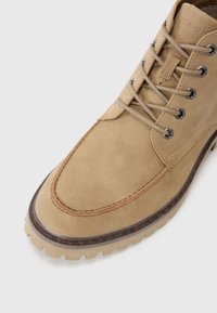 Bota de ante marrón claro con punta redondeada, detalles cosidos, ojales de metal y una suela de goma resistente con surcos profundos para tracción.