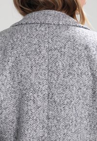 Šedý kabát ze směsi vlny s texturovaným povrchem, klasickým límcem a tvarovaným zadním švem. Hladký povrch s jemnými barevnými variacemi.