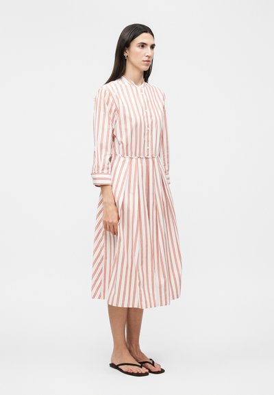 Γυναίκα που στέκεται πλαγίως φορώντας λευκό φόρεμα μεσαίου μήκους με κάθετες πορτοκαλί ρίγες και μαύρες σαγιονάρες σε μονόχρωμο φόντο.