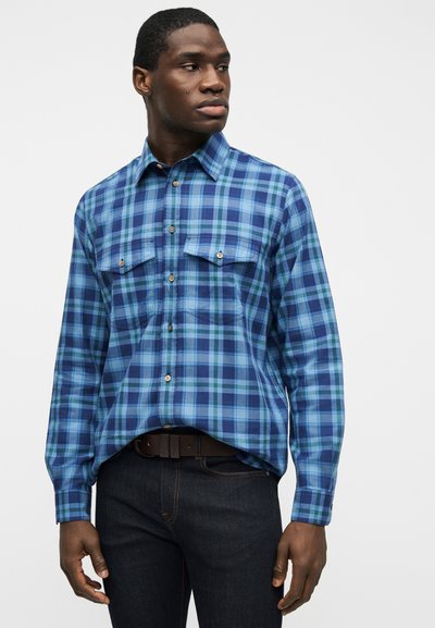 Άνδρας που φοράει μακρυμάνικο πουκάμισο με κουμπιά σε μπλε και τιρκουάζ ψαροκόκκαλο και σκούρο τζιν, κοιτάζοντας προς το πλάι με φόντο έναν απλό τοίχο.