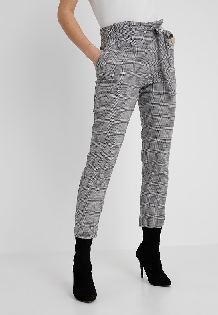 Femme portant un pantalon paperbag taille haute à carreaux gris avec ceinture nouée et des bottines noires à talons hauts, debout les mains dans les poches.