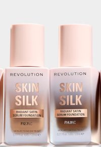 Två flaskor med Revolution Skin Silk Radiant Satin Serum Foundation. Rosa lock, klara behållare, etiketter med guldtext som anger nyansnummer F12.7NC och F14.9NC.