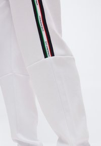 Λευκό παντελόνι με μαύρη, πράσινη, λευκή και κόκκινη κάθετη ρίγα στο πλάι με τη σήμανση EA7 Emporio Armani.