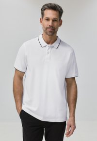 Homme portant un polo blanc avec une bordure noire et un pantalon noir, debout avec une main dans la poche devant un fond clair uni.