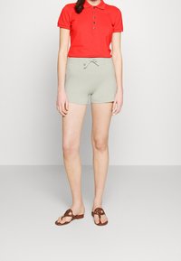 Kvinna som bär en röd kortärmad polo, ljusgrå stickade shorts med en rosett och bruna öppna sandaler med slip-on, stående mot en enkel bakgrund.