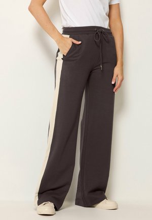 Personne porte un t-shirt blanc et un pantalon ample foncé avec des bandes latérales beiges, taille à cordon et baskets beiges, debout avec une main dans la poche.