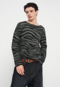 Γκρι πουλόβερ με μαύρο κυματοειδές σχέδιο, κατασκευασμένο από μαλακό ύφασμα, διαθέτει φαρδιά λαιμόκοψη και μακριά μανίκια, συνδυασμένο με σκούρο τζιν και καφέ ζώνη.