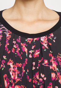 Schwarze Bluse mit floralem Muster in lebhaften Pink- und Rottönen, gefaltetem Design und einem schwarzen gerippten Ausschnitt. Weicher Stoff.