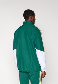 Vihreä urheilutakki, jossa valkoiset ja kultaiset korostukset, korkea kaulus ja joustava vyötärö. Mallissa on väriblokatut hihat ja urheilulliset raidat.