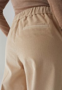 Pantaloni in corduroy beige con finitura testurizzata, dotati di una cintura elastica e di una singola tasca posteriore. Dettagli in tessuto a coste morbido visibili.