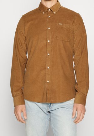 Mann trägt ein braunes Cordhemd mit langen Ärmeln und Brusttasche, kombiniert mit hellblauen Jeans, steht vor einem schlichten Hintergrund.