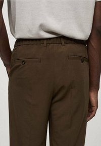 Braune, maßgeschneiderte Hose mit elastischem Bund, zwei Gesäßtaschen mit Knopfverschlüssen und glatter Textur; Kleidungsstück von hinten betrachtet.