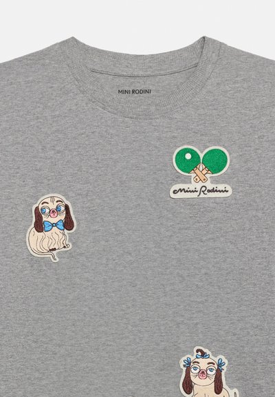 Γκρι πουκάμισο με κεντημένα patches δύο σκύλων που φορούν γυαλιά και παπιγιόν, και δύο πράσινα ρακέτες πινγκ πονγκ με το κείμενο "Mini Rodini".