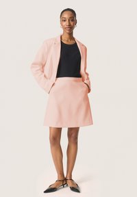 Rausvaišis švarkas su juodu ribotu topu ir suderintomis sijonais. Juodi smailūs butai su kulkšnies dirželiais. Lygus audinio paviršius.