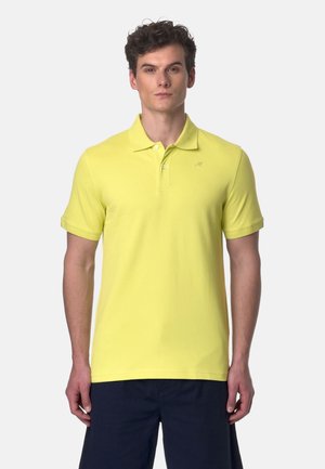 Uomo che indossa una polo gialla vivace a maniche corte e pantaloncini blu scuro in piedi davanti a uno sfondo chiaro e uniforme.