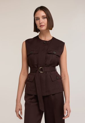 Frau mit geradem Bob-Haarschnitt, die eine ärmellose dunkelbraune, taillierte Weste mit geknöpften Klappentaschen und passenden Hosen trägt, stehend vor neutralem Hintergrund.