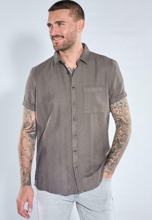 Mann mit kurzen Haaren und Tattoos auf beiden Armen, trägt ein graues kurzärmeliges Hemd und helle Jeans, lächelt und schaut nach links.
