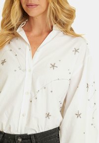 Camicia bianca con bottoni, a maniche lunghe, con dettagli argentati e cuciture che ricordano stelle, abbinata a jeans scuri.