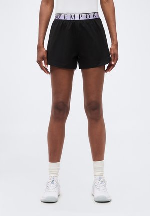 Person trägt schwarze Sportshorts mit einem violetten Emporio Armani-Bund, weiße Socken und weiße Sneakers, steht vor einem schlichten Hintergrund.