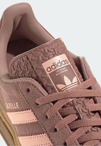 Baskets en suède marron avec des accents texturés roses, arborant un logo à trois bandes, des lacets épais et une conception à bout rond.