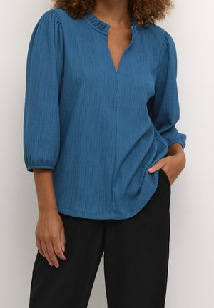 Vrouw draagt een blauw textuurblouse met 3/4 pofmouwen en een kleine geplooide kraag, gecombineerd met zwarte broek, hand in de zak.