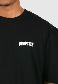 Sort bomulds t-shirt med et hvidt "DROPSIZE" logo i en fed, stiliseret skrifttype på venstre brystområde. Korte ærmer, rund halsudskæring.