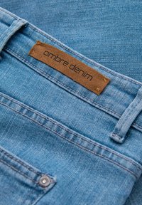 Světle modrý denimový materiál s hnědým semišovým štítkem přišitým blízko poutka na opasek s nápisem "ombre denim."