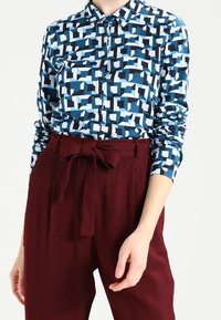Long-sleeve button-up shirt in blue tones with a geometric pattern; paired with high-waisted burgundy pants featuring a tied belt.