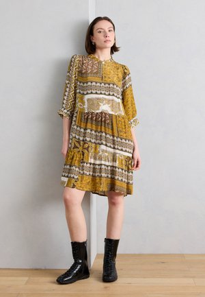 Nő áll egy fal mellett, sárga és barna mintás, térdig érő ruhát visel 3/4-es ujjal és fekete fűzős csizmával.