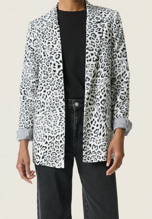 Femme portant un blazer blanc et noir à imprimé léopard sur une chemise noire et un jean noir taille haute, debout les mains détendues le long du corps.