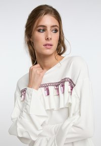 Weißes Langarmtop mit Rüschenärmeln, verziert mit einem dekorativen rosa Quastenbesatz und Kettendetails über der Brust. Glatte Stofftextur.