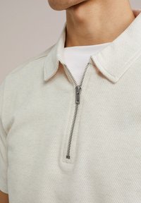 Bela, teksturirana polo majica na zips s ovratnikom in srebrnim zadrgo. Pod njo je navadna bela majica, ki prikazuje gladek kontrast tkanine.