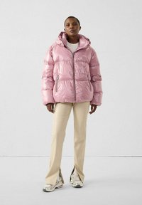 Roze pufferjack met een glanzende afwerking, capuchon, rits aan de voorkant en zijzakken. Draagt over lichtbeige broek en neutrale sneakers.