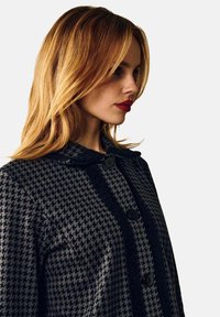 Sort og grå frakke med houndstooth-mønster, krave, sorte knapper og struktureret materiale, vist fra siden.