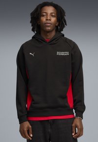 Sort hættetrøje med røde sidepaneler, der har en hætte, frontkangaroo-lomme og "SCUDERIA FERRARI" logo i hvidt på brystet.
