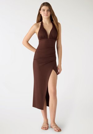 Donna in piedi che indossa un vestito marrone senza maniche con scollo all'americana e una spacco alto, abbinato a sandali piatti beige con cinturini, sfondo neutro.