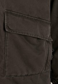 Schwarzer Baumwollstoff mit einer großen aufgenähten Tasche, detaillierten Nähten und einer leicht strukturierten Oberfläche. Einfaches Design ohne sichtbare Muster.