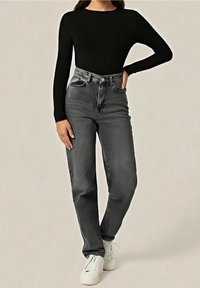 Femme portant un haut noir cintré à manches longues, un jean gris taille haute et des baskets blanches à lacets, debout avec une main sur la hanche.