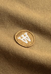 Rudos audinys su tekstūruota paviršiaus struktūra turi apvalų lopelį. Lopelyje yra auksinė fono spalva ir baltos siuvinėtos raidės „WW“.