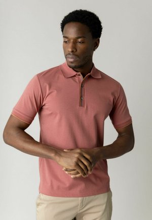 Uomo con polo rosa polvere e pantaloni beige in piedi con le mani incrociate, guarda verso il basso e di lato su uno sfondo semplice.