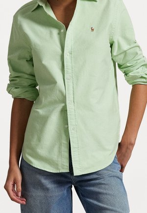 Personne portant une chemise boutonnée vert clair à manches retroussées et un jean bleu, debout avec une main dans la poche et l'autre détendue.